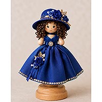 Handmade doll pin doll featuring a royal blue dress, matching floral hat, curly brown hair, and decorative ribbon rose embellishments displayed on a wooden stand.